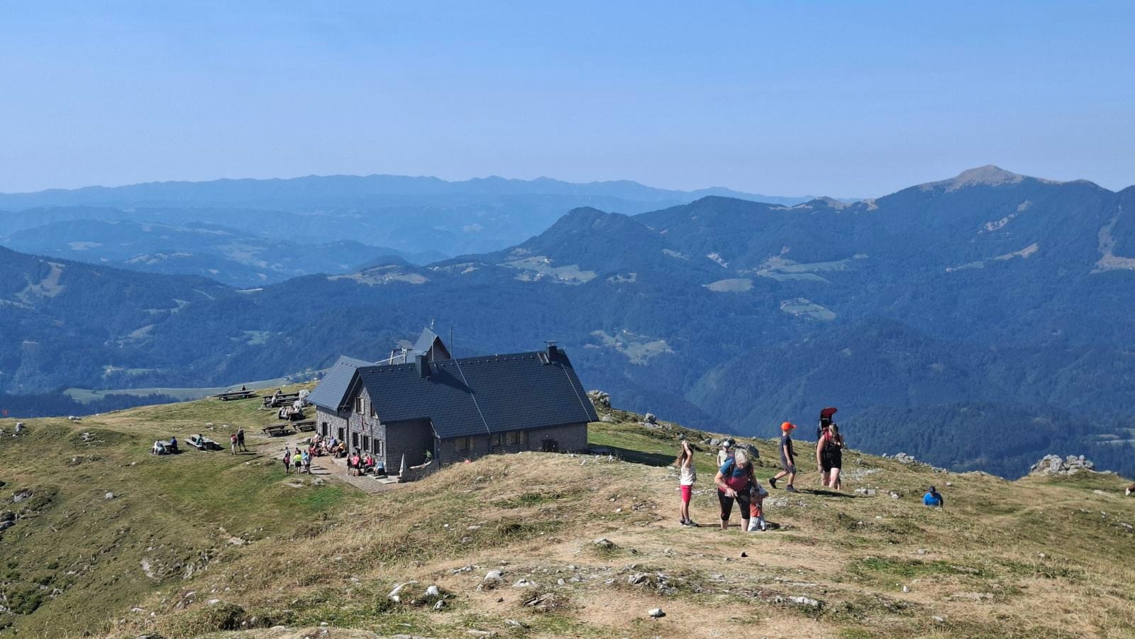 Krekova koča na Ratitovcu with hikers relaxing on the grassy ridge outside, surrounded by rolling hills